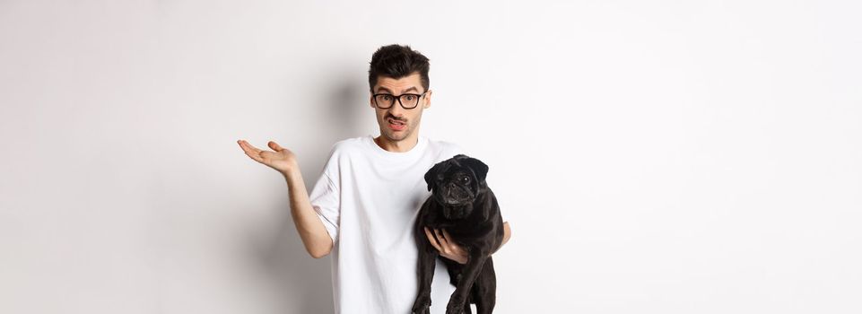 Image of handsome young man holding black pug and looking confused. Guy shrugging shoulders and staring indecisive at camera, carry dog in arm, white background.