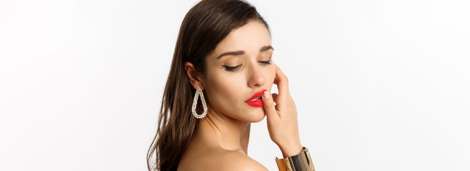 Fashion and beauty concept. Tender and feminine brunette woman touching red lips and looking down sensually, standing over white background.