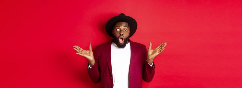 Fashion and party concept. Impressed Black man in classy outfit staring with complete disbelief at camera, gasping and looking surprised, standing over red background.