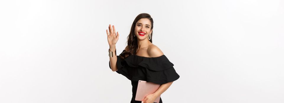 Fashion and shopping concept. Stylish young woman with glamour makeup, wearing black dress, holding digital tablet and saying hi, waving hand to greet you, white background.