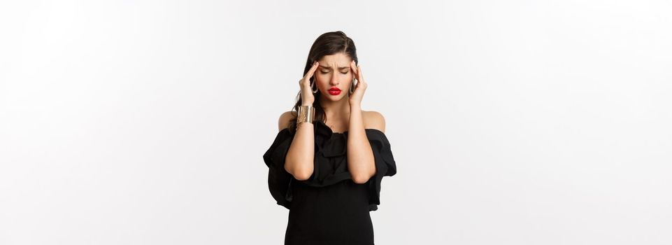 Fashion and beauty. Young modern woman in black dress, red lipstick, having headache, touching head and feeling sick, standing over white background.