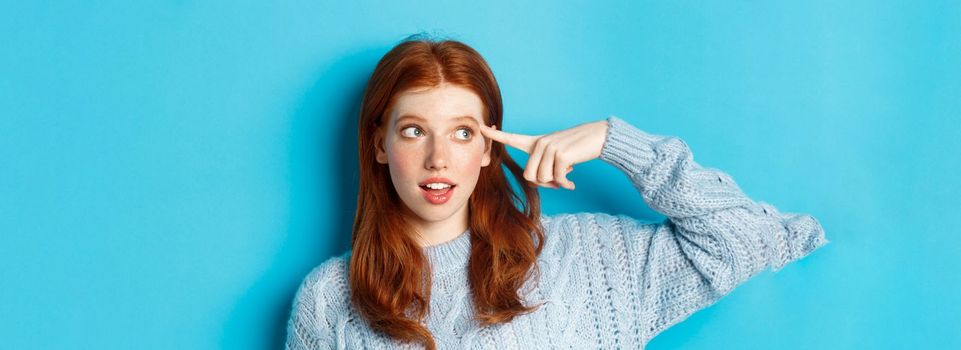 Close-up of cute redhead girl searching for solution, pointing at head and looking left at logo, standing over blue background.