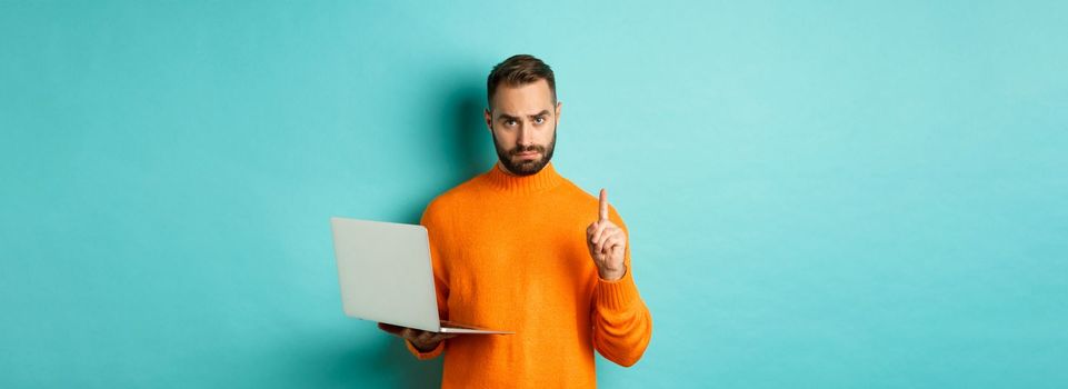 Image of adult man teaching online, showing finger while using laptop, explaining rules, standing over light blue background.