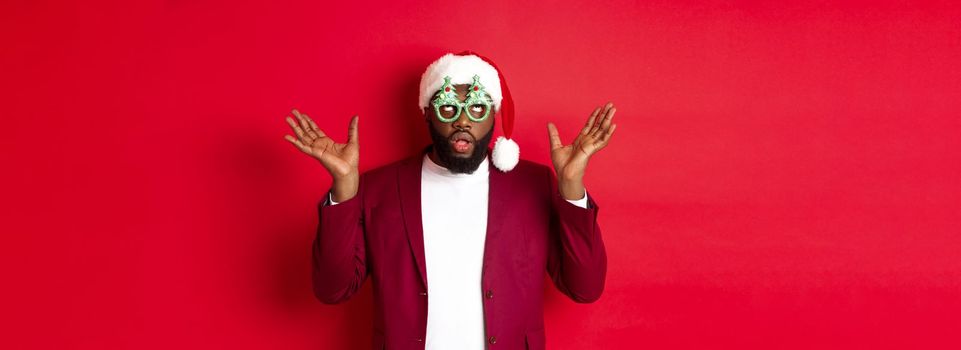 Merry Christmas. Cheerful Black man wearing funny party glasses and santa hat, smiling joyful, celebrating winter holidays, standing over red background.