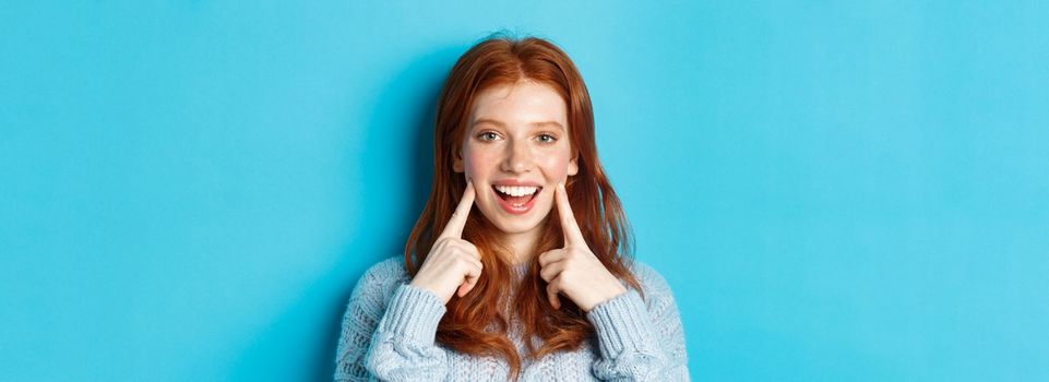 Close-up of cheerful teenage girl with red hair and freckles, poking cheeks, showing dimples and smiling with white teeth, standing over blue background.