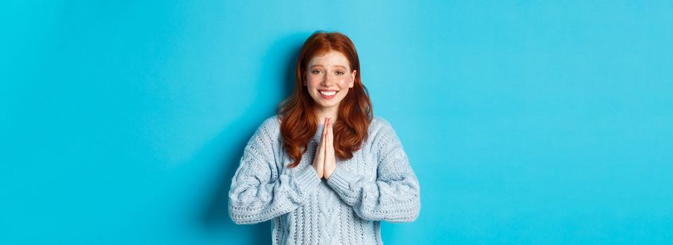 Cute redhead girl saying thank you, smiling and looking at camera, expressing gratitude, standing against blue background.