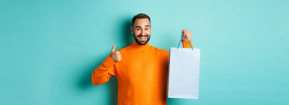 Handsome smiling man showing thumbs-up and shopping bag, recommending store, standing over blue background. Copy space