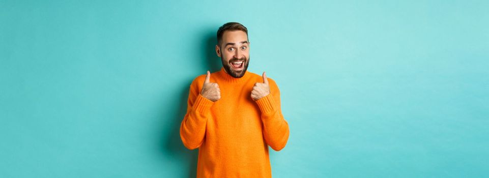 Excited young man with beard, showing thumbs up in approval, praise or recommend, standing over light blue background.