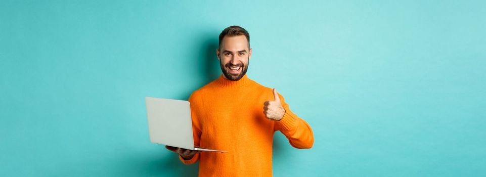 Freelance and technology concept. Lucky man in orange sweater, showing thumb up while working with laptop, standing satisfied over light blue background.