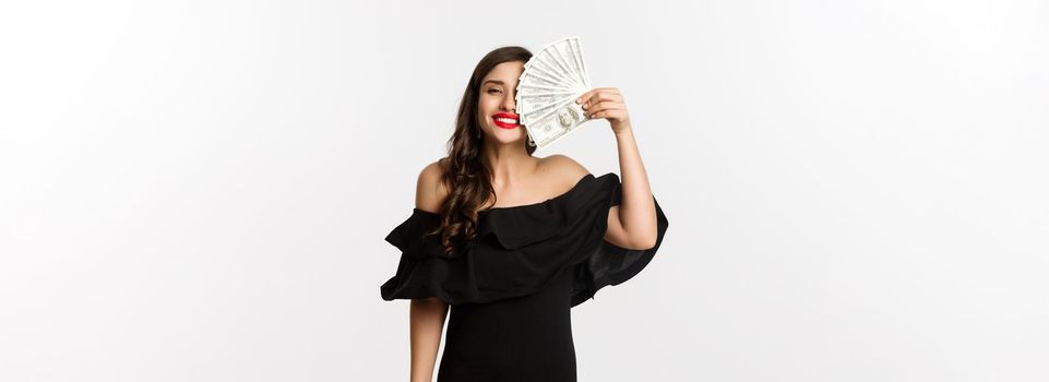 Fashion and shopping concept. Happy young woman in black dress, with red lips, holding money and smiling satisfied, standing over white background.