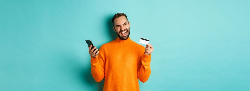 Online shopping. Happy young man using mobile phone and credit card, paying internet, light blue background.