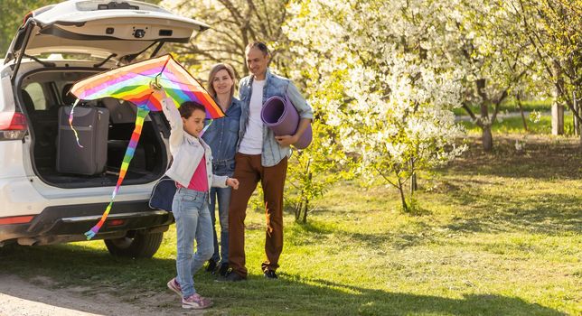 Happy family standing together near a car with open trunk enjoying view of rural landscape nature. Parents and their kid leaning on vehicle luggage compartment. Weekend travel and holidays concept