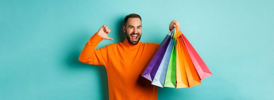 Handsome bearded man in orange sweater, buying gifts, pointing at himself and showing shopping bags, standing over blue background.