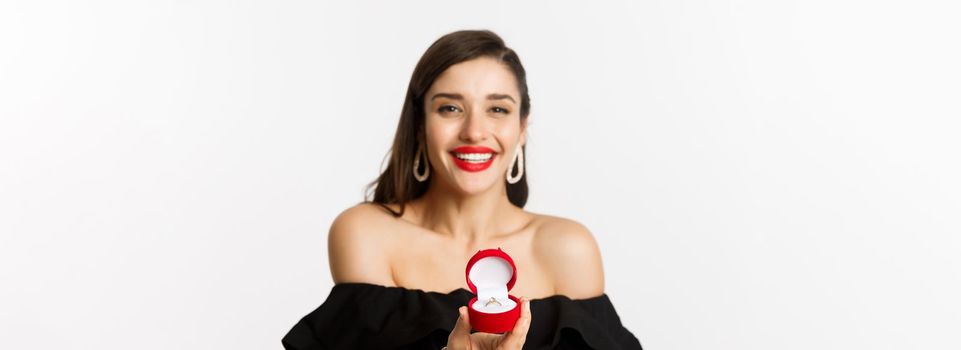 Fashion and beauty concept. Close-up of gorgeous brunette woman with red lips, black dress, showing engagement ring in box and smiling, getting married, standing over white background.