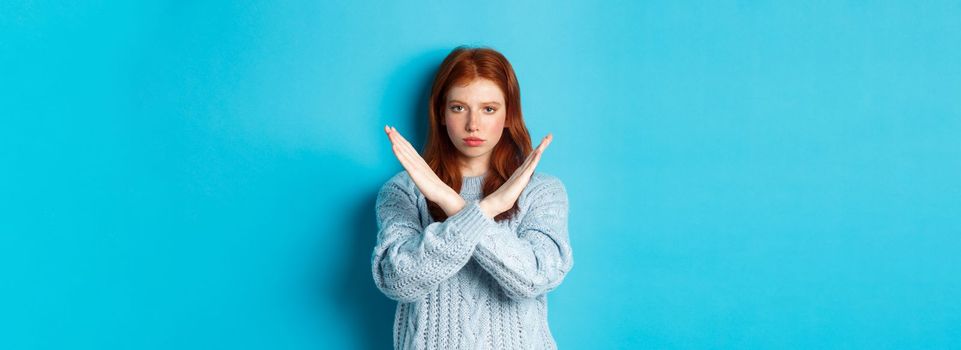 Serious redhead girl looking confident, showing cross gesture to stop and forbid action, standing over blue background.