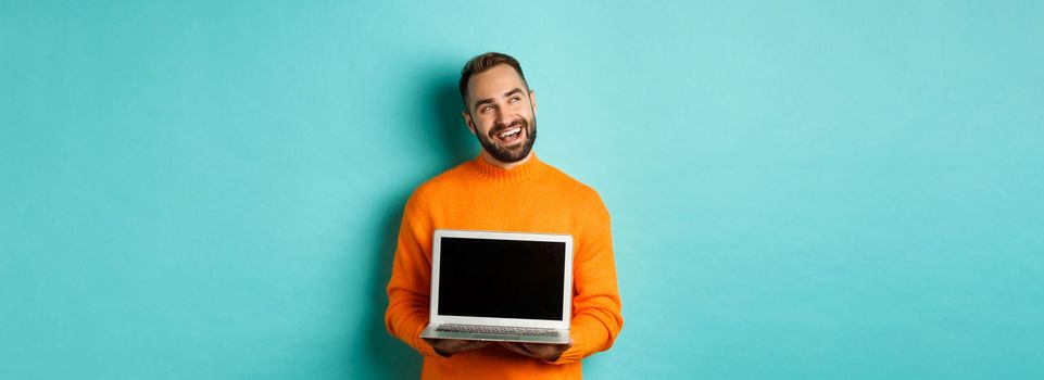 Happy caucasian man thinking and showing laptop screen, smiling pleased while pondering, standing in orange sweater against light blue background.