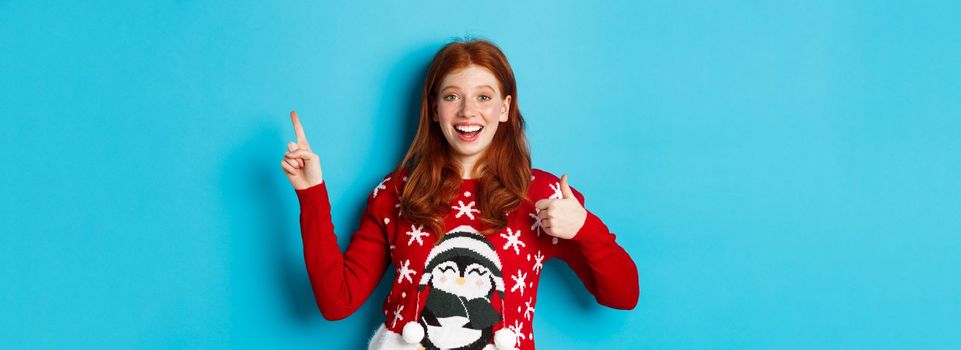 Merry Christmas. Cheerful redhead girl in xmas sweater, pointing finger at upper right corner, showing new year promo and thumbs-up in approval, praise product.