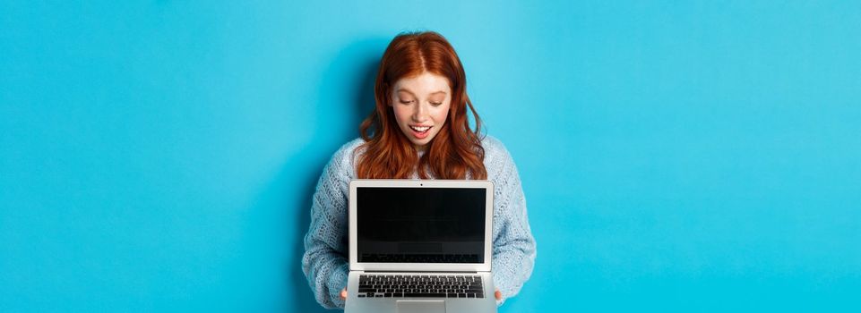 Amazed redhead girl staring at laptop screen and looking impressed, showing computer display, standing over blue background.