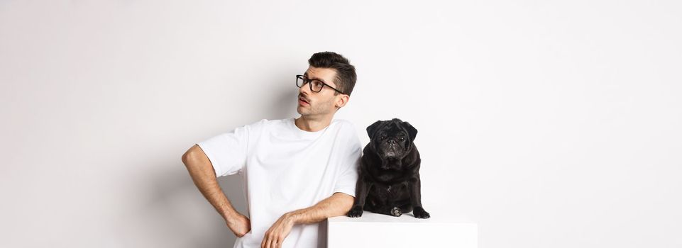 Handsome young man standing near cute black pug, looking right with arrogant expression, standing over white background.