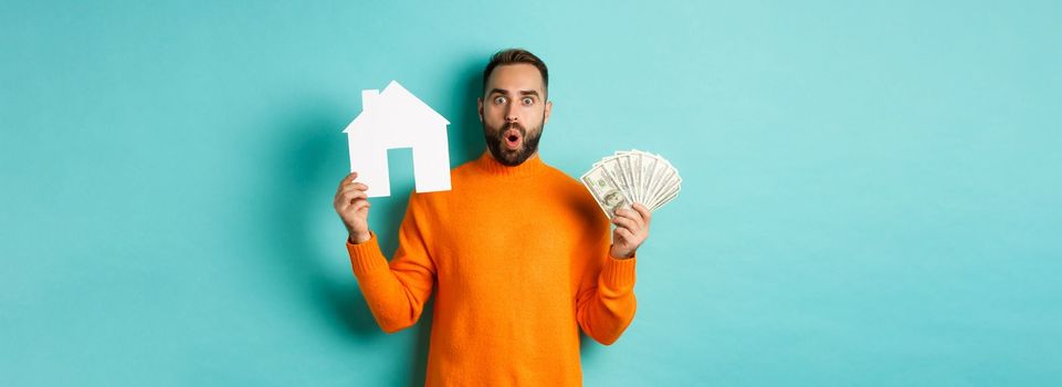 Real estate concept and mortgage concept. Amazed buyer holding money and paper house maket, buying apartment, standing over light blue background.