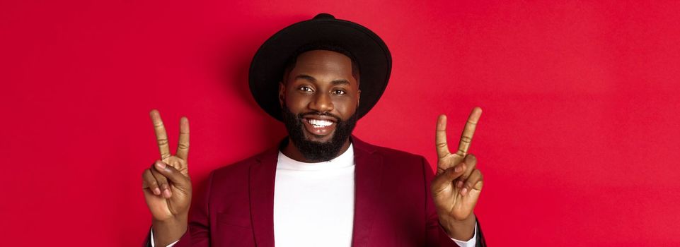 Close-up of handsome bearded Black man showing peace signs and smiling happy, posing for phoro, standing over red background.