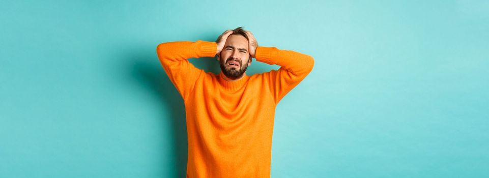 Image of frustrated man in orange sweater, holding hands on head and feeling distressed, standing anxious against turquoise background.