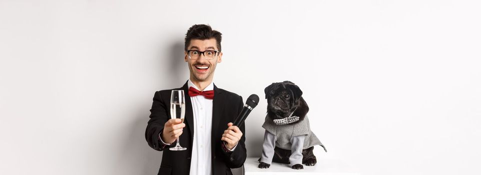 Happy guy raising glass of champagne, giving microphone to dog in costume, celebrating and enjoying party, standing over white background.