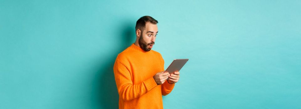 Image of male model in orange sweater staring at digital tablet screen, looking surprised, standing over light blue background.