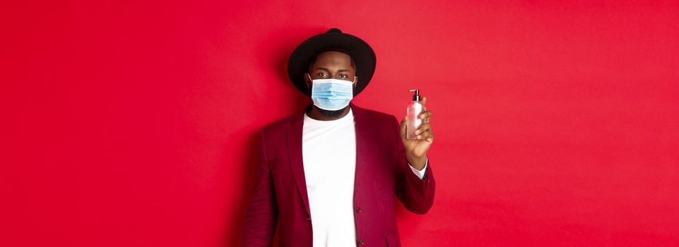 Covid-19, quarantine and holidays concept. Young african american man in medical mask and stylish outfit showing hand sanitizer, recommending to use antiseptic.