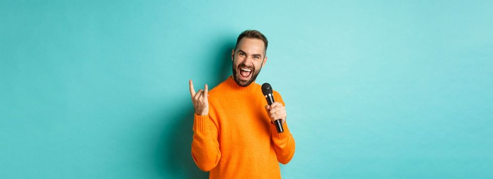 Joyful handsome man singing song in microphone, showing rock-n-roll gesture excited, standing over turquoise background.