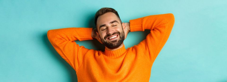 Carefree young man daydreaming, resting with hands behind head and closed eyes, relaxing over turquoise background in orange sweater.