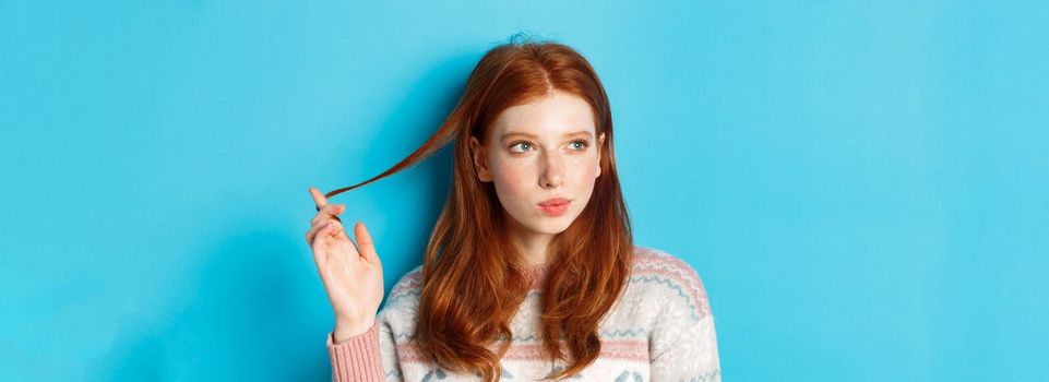 Close-up of thoughtful pretty redhead girl looking left, playing with hair strand and pondering, standing in winter sweater against blue background.