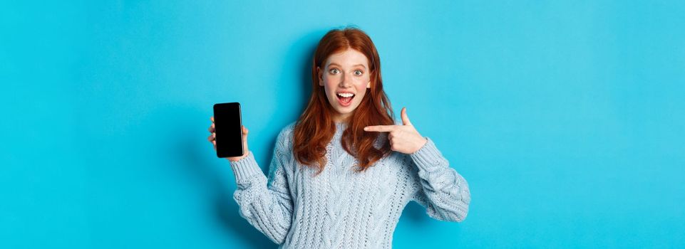 Impressed redhead girl pointing at phone screen, showing smartphone app or online offer and smiling excited, standing in sweater against blue background.