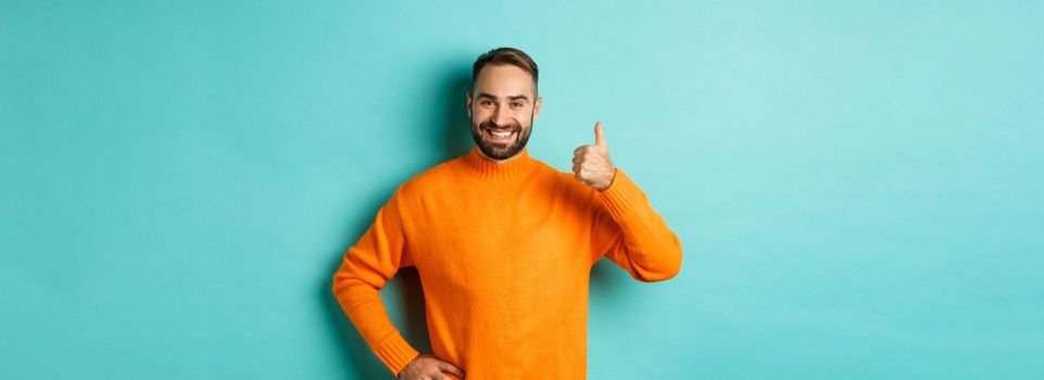 Well done. Handsome bearded man showing thumb up, praising good work, recommend excellent product, like and approve, smiling satisfied, standing over light blue background.