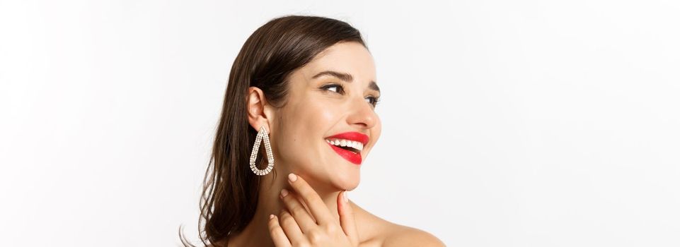 Fashion and beauty concept. Headshot of gorgeous brunette woman with red lipstick, earrings, laughing and looking left, standing over white background.