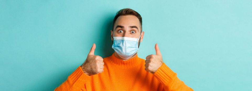 Covid-19, social distancing and quarantine concept. Close-up of satisfied male model in medical mask showing thumbs-up, praising and agreeing, standing over light blue background.