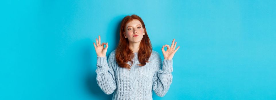 Redhead teenage girl showing okay signs, looking satisfied and proud, agree, give positive answer, praising good choice, standing over blue background.