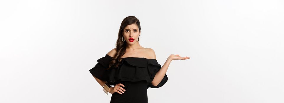 Fashion and beauty. Annoyed woman in black dress raising hand, so what gesture, looking confused at camera, standing bothered over white background.