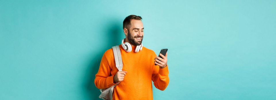 Caucasian man with headphones and backpack looking at phone, reading message and smiling, standing over turquoise background.