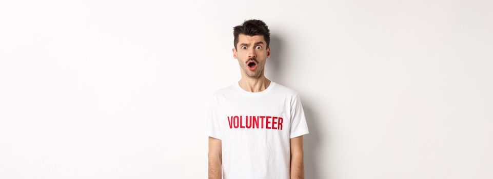 Shocked and confused man in volunteer t-shirt staring at camera speechless, standing against white background.