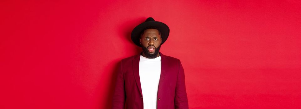 Fashion and party concept. Confused Black man in classy outfit frowning and staring at camera displeased, cant understand, standing over red background.