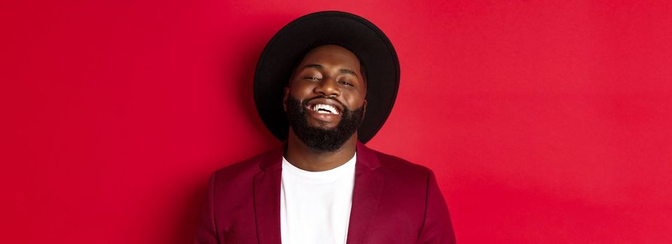 Close-up of happy handsome Black man smiling at you, looking pleased, wearing black hat and blazer, red background.