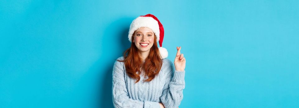 Winter holidays and Christmas Eve concept. Hopeful redhead girl in Santa hat, making wish on xmas, cross fingers for good luck and smiling, standing over blue background.