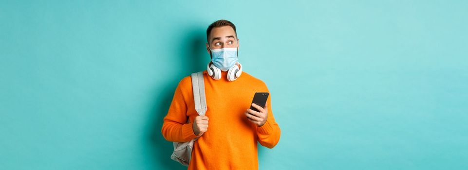 Young man in face mask using mobile phone, holding backpack, staring left amazed, standing against light blue background.