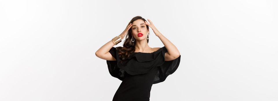 Confident and stylish woman in black elegant dress, looking sassy at camera, standing over white background.