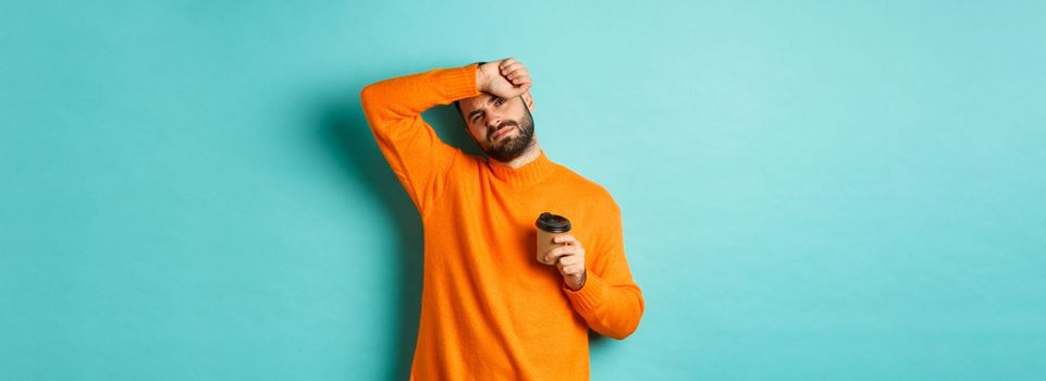 Tired adult man taking break from work and drinking coffee, wipe sweat off forehead and looking exhausted, standing in orange sweater over turquoise background.