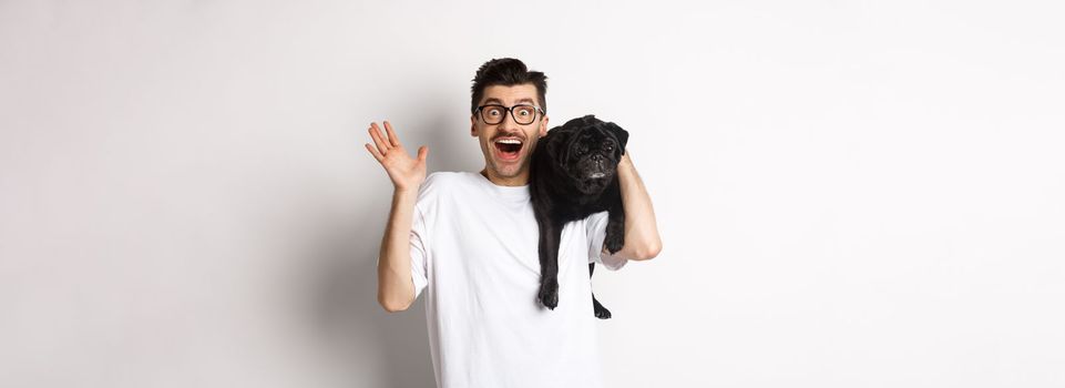 Excited hipster guy in glasses holding dog on shoulder, waving hand to say hello, greeting someone with happy face. Happy man carry his cute pug and smiling, white background.