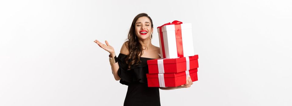 Celebration and christmas holidays concept. Excited and happy woman receive gifts, holding xmas presents and rejoicing, standing in black dress over white background.