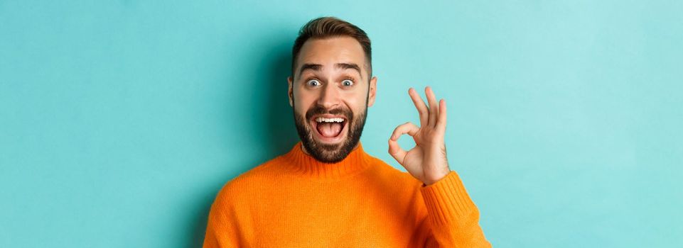 Impressed handsome man showing okay sign, recommending something good, praising with ok gesture, standing over light blue background.