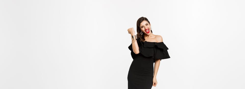 Beauty and fashion concept. Full length shot of happy young woman in luxury black dress, jewelry and heels, wearing makeup and looking satisfied, celebrating victory, winning, white background.